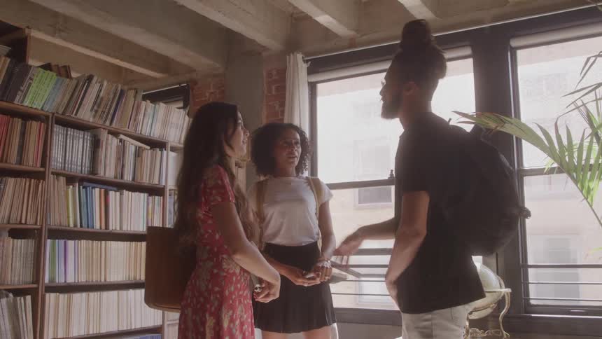 Three people talk in a bright room with a bookshelf full of books