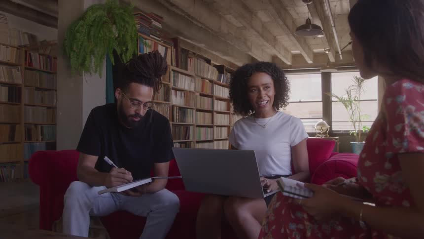 Three people are gathered around a coffee table one person sits and writes in a book another person sits with a laptop and talks and another person listen to them