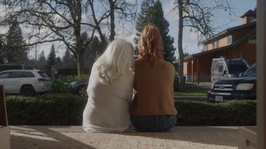 Two women sit with their backs to the camera on a porch looking out at a street in a suburb setting with cars and other houses