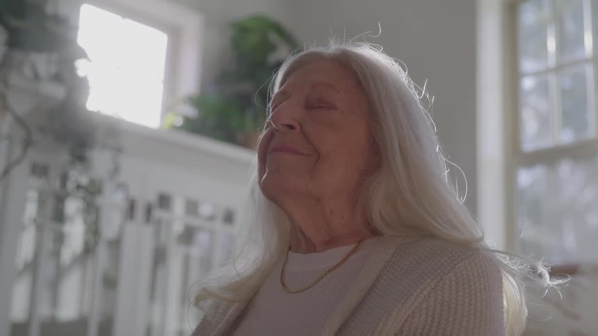 A woman with her eyes closed is in a room with windows and potted plants behind her