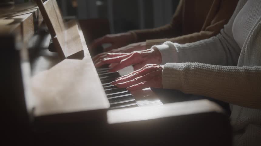 A close view of the hands of an elderly woman and a younger woman playing the keys of a wooden upright piano