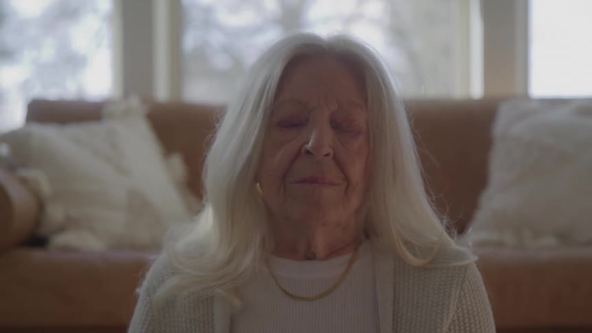 An elderly woman sitting on the floor with her eyes closed on an indoor setting