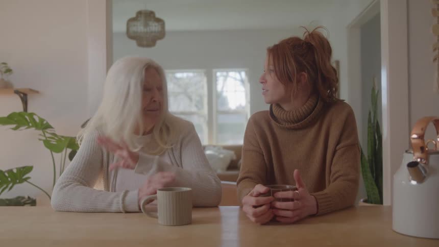 An elderly woman with white hair and a younger woman with reddish hair are sitting at a bright kitchen counter with mugs talking and gesturing to each other