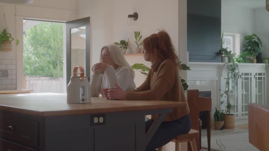 Two people sit facing each other at a kitchen island and talk while holding mugs