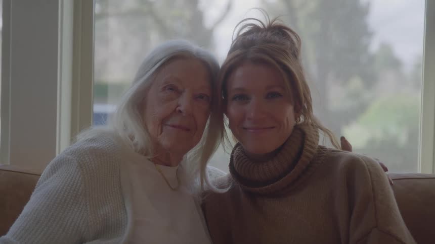 Two woman sit side by side on a couch and the elderly woman has one arm around the other woman and both are looking at the camera