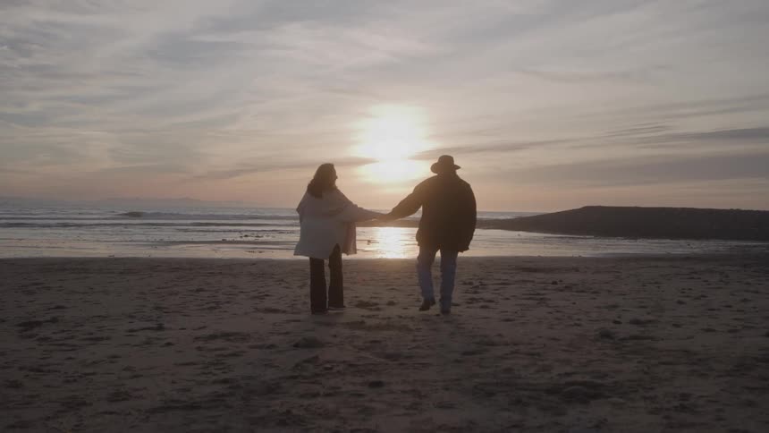 Couple holding hands walking on beach towards shoreline at sunset