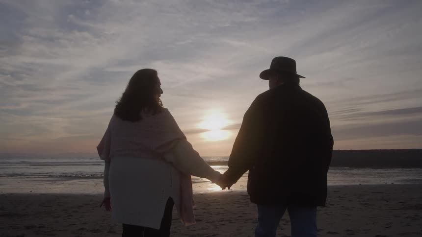 A man wearing a cowboy hat holding hands with a woman as they walk together on the beach sand at sunset