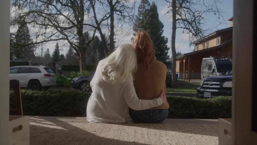 Whitehaired woman hugs a young woman sitting beside her on the porch
