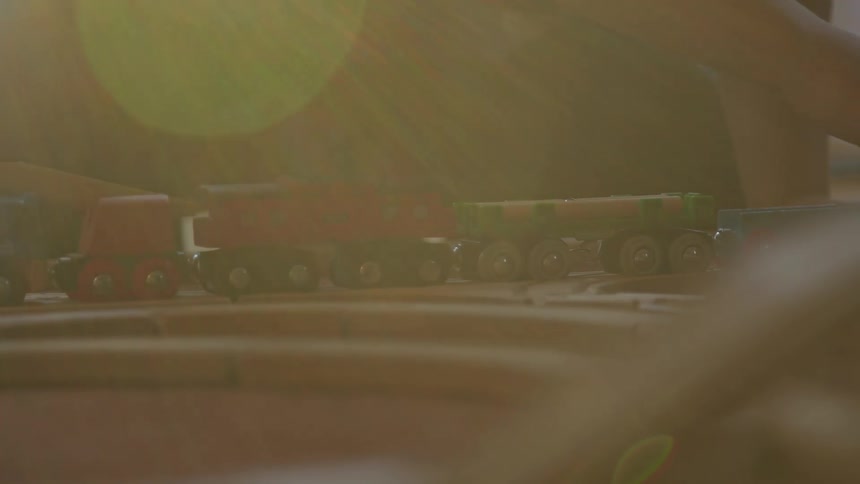 Childs hand moves toy train along wooden tracks with colorful cars lit by sunlight