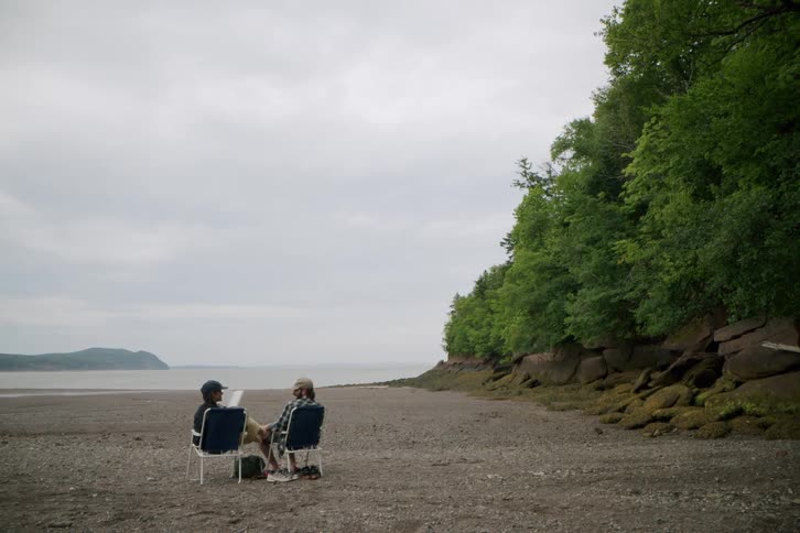Two women sitting on chairs on a beach positioned far from the shoreline engaged in a lively and fun conversation at midday trees border the right side of the scene