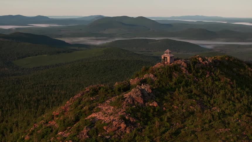 A tower stands on top of a hill and is surrounded by mountains and trees with a mistfilled valley behind it