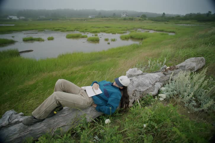 A young woman is lying on a tree trunk in a lake setting with a book on her lap