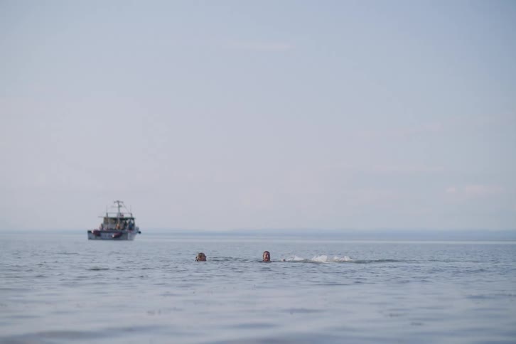 Person playing in the water while another swims nearby and a boat appears in the background