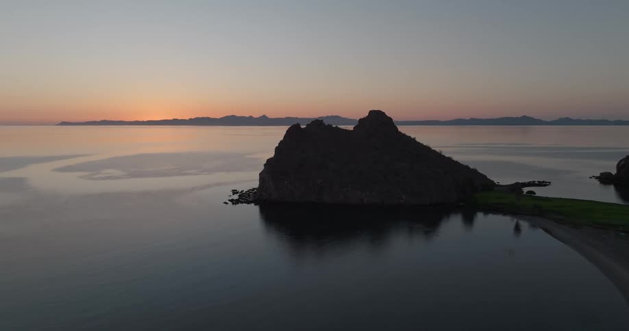 Coastal rock formation in silhouette contrasts with colorful sunset sky reflected on calm waters before mountain range in the background