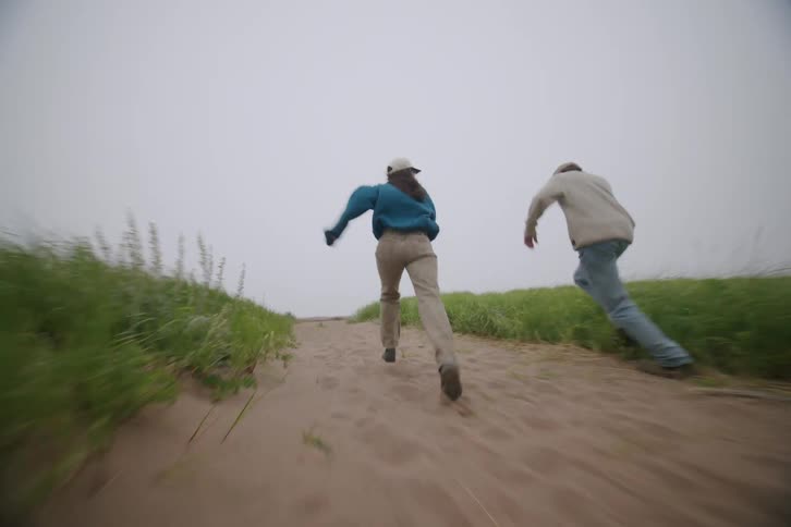 A man and a woman in casual clothes run along a sandy path with tall grass on either side in a foggy outdoor environment