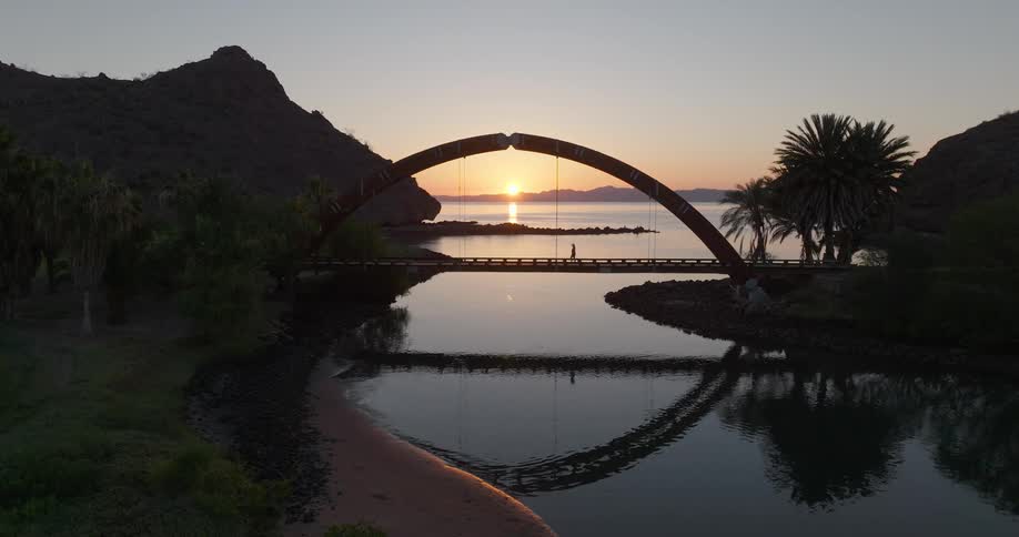 Person walks across arched bridge during sunset with symmetrical reflection on water between palm trees and rocky hills