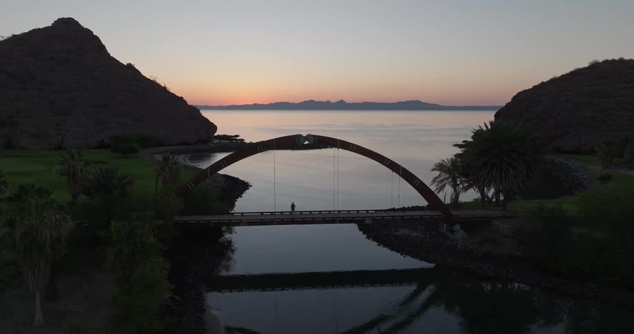 A person walks across a bridge over a body of water at sunset
