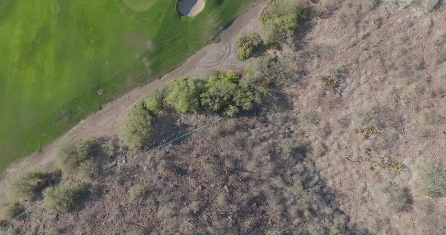 An overhead shot of arid rocky terrain scattered with sparse scrub and leafless bushes then there are lush grassy area next to clear ocean