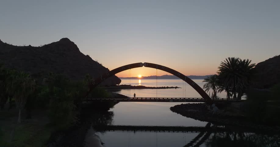 Arched bridge with one person standing on top frames the sunset reflected on water between mountain and palm trees