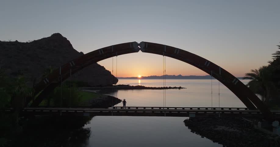 A bridge over water with a sunset or sunrise over the horizon a person walks on the bridge toward the camera