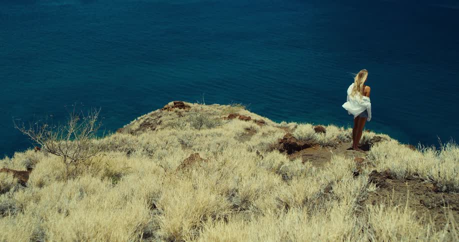 A woman with long blonde hair and a white shirt stands on a cliffside looking out at the dark blue water below the cliff is covered in dry goldencolored grass and small rocks