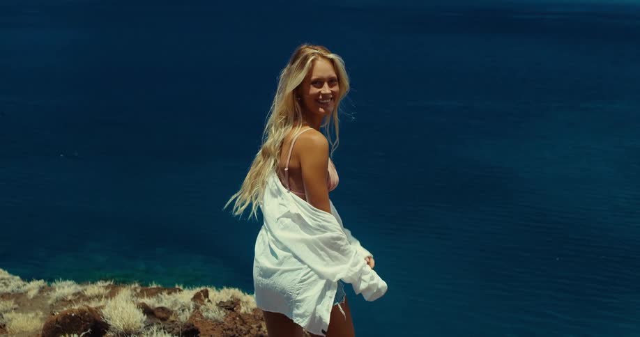 Smiling blonde woman turns from facing the sea and walks away from the edge of the shore on a summer day