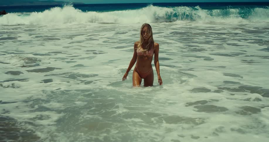 A wide shot captures a woman with long blonde hair and a white bikini standing in the ocean the waves crash around her as she smiles seemingly enjoying her day at the beach
