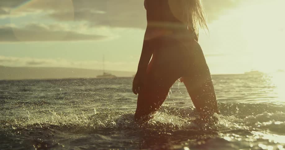 Blonde woman wading into the surf is momentarily silhouetted by the sun sitting on the horizon