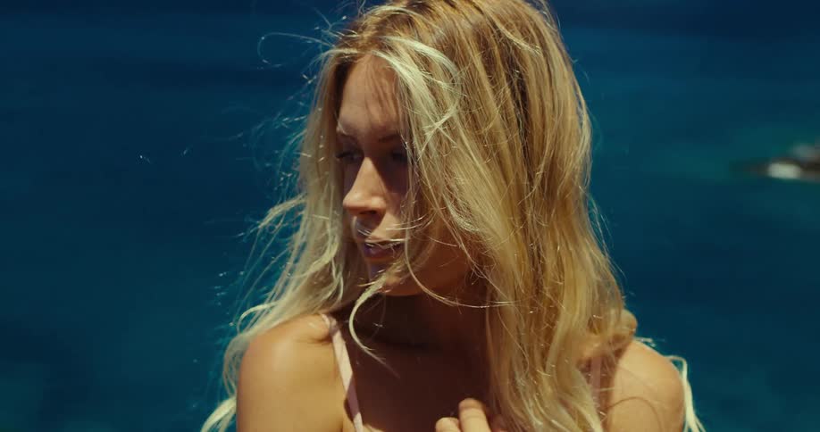 Blonde woman with loose hair looks to the side in front of the deep blue sea under bright light