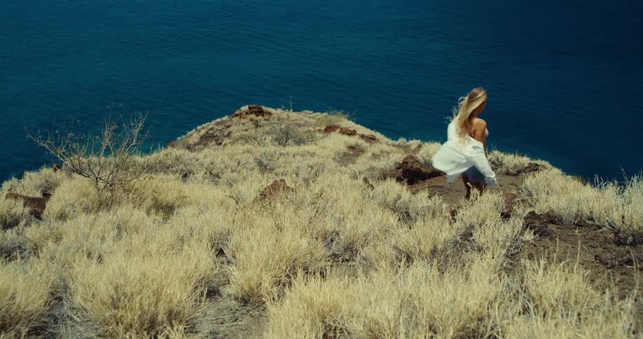 Blonde woman walks on a dry hillside overlooking the blue sea