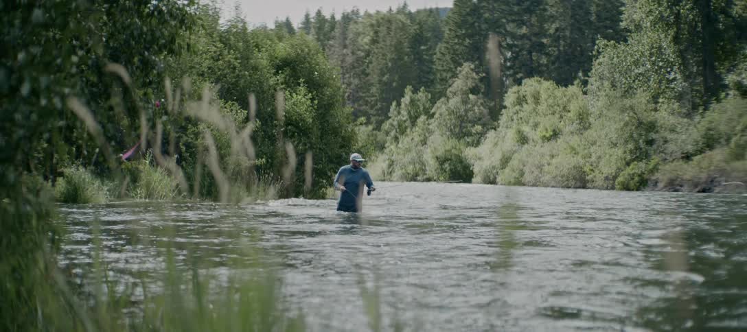 A man wearing waders and a hat is standing in the middle of a flowing river fly fishing the river is surrounded by dense greenery and trees