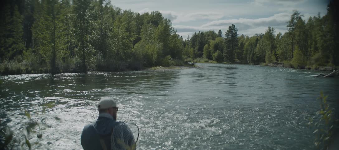A man wearing a hat and a vest stands on the bank of a river he is holding a fishing net looking out at the water surrounded by a lush green forest