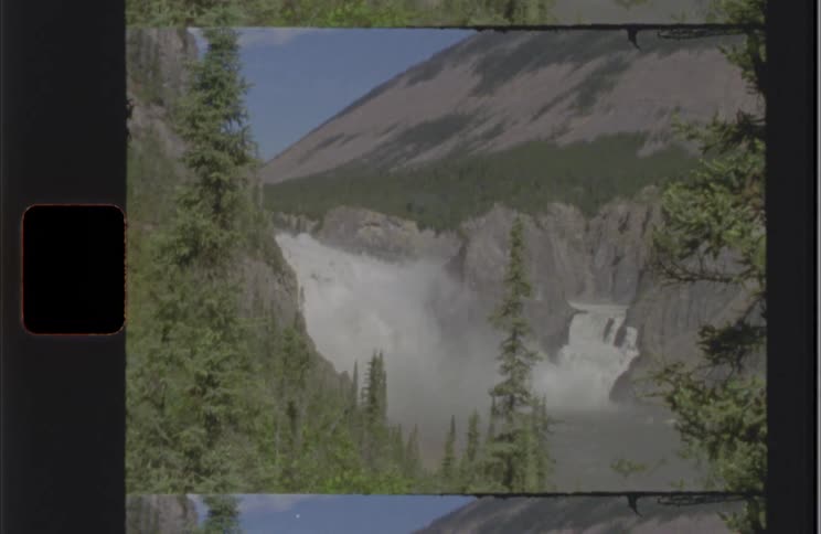 A waterfall cascades amid green trees and rocks under a blue sky