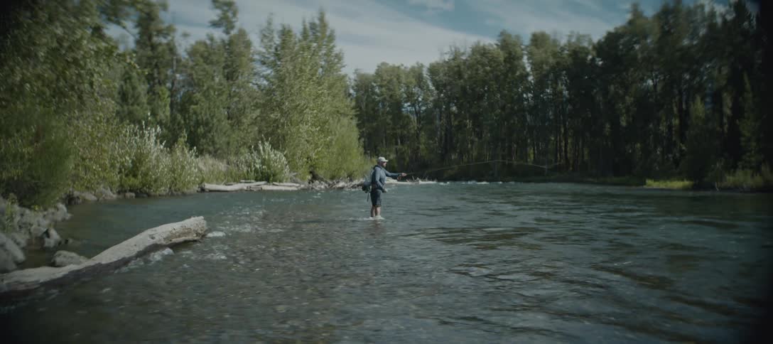 Angler flyfishing in a flowing river surrounded by trees in the wilderness
