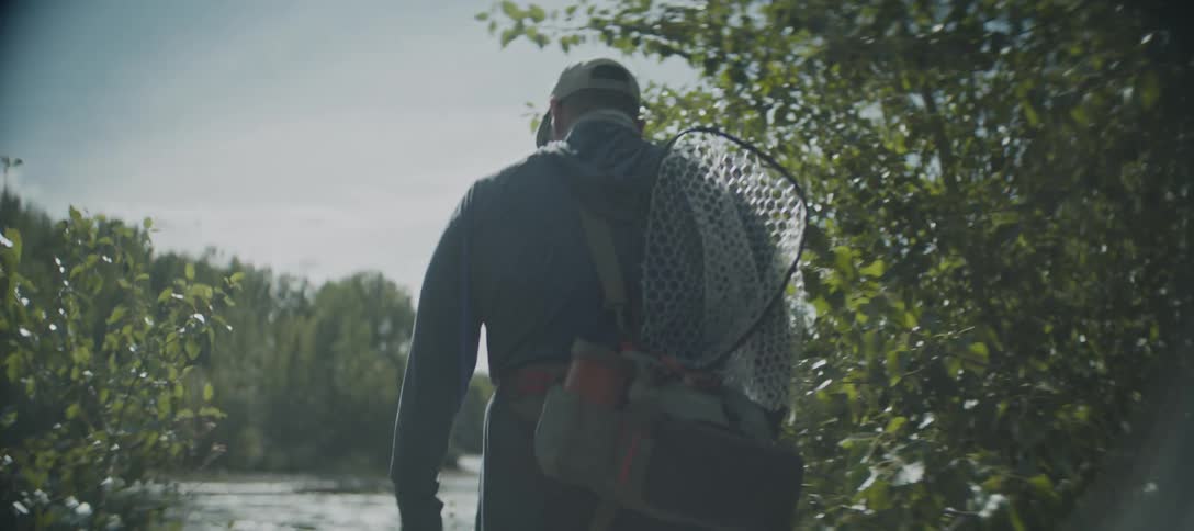 A man in a baseball cap and dark blue jacket carrying a fishing net and gear on a strap walks through sunlit green trees to the rivers edge