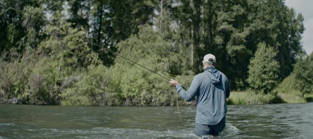 Person in fishing attire casts line into river surrounded by dense vegetation and moderate current