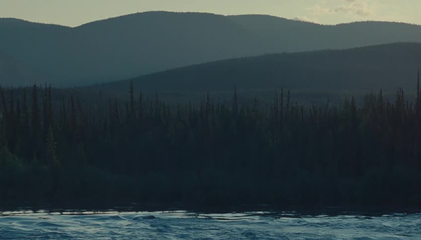 Coniferous forest stretches behind gently rippling river with layered mountains in the background