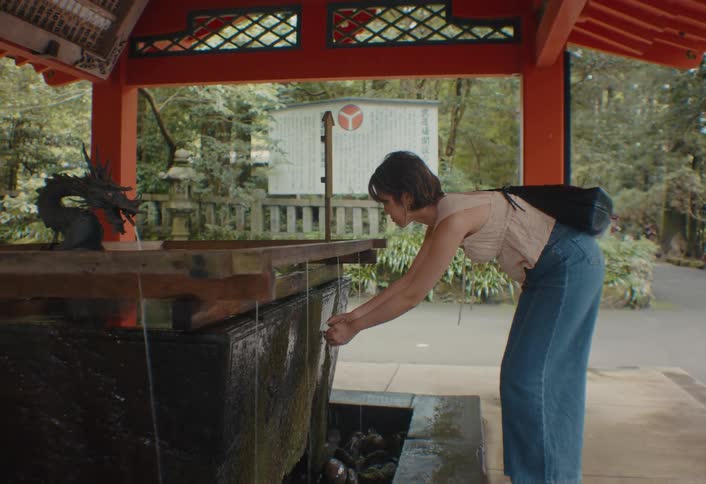 A young woman wearing jeans and holding a handbag washes her hands at a public tap outdoors at midday