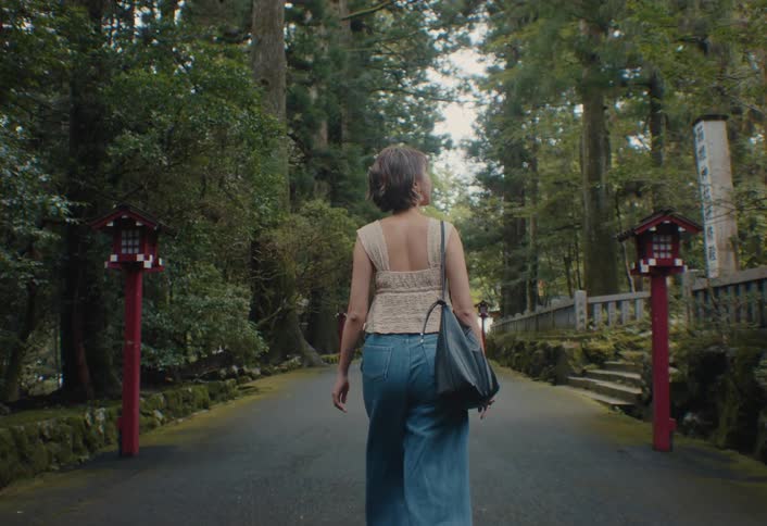A woman with short hair and a black bag walks away from the camera down a path lined with tall trees and traditional japanese lanterns