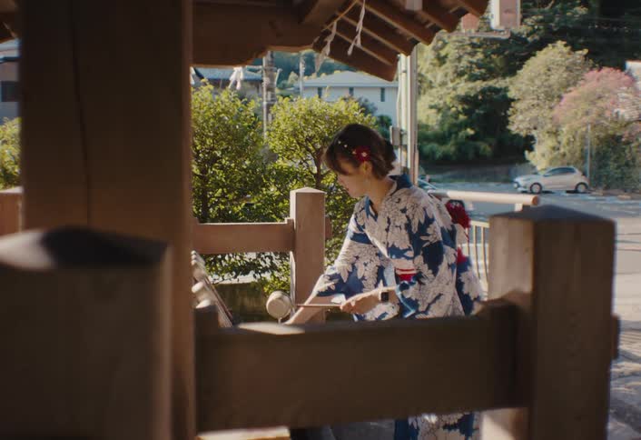 A young woman in a blue and white patterned yukata with a red obi is performing a ritual water purification at a wooden shrine on a sunny day