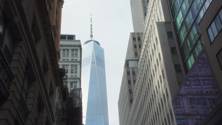 A camera captures a vertical view from a street framed by tall older buildings on the sides focused on a monumental glassclad tower rising into the sky