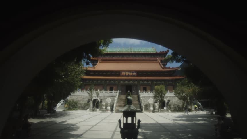 A large chinese temple building with red and gold eaves and a stone staircase is viewed through a dark archway centered around a bronze censer in a sunny courtyard