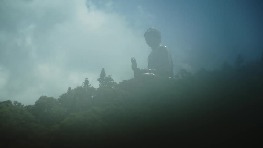 A large statue of a seated buddha is visible on a mountaintop partially shrouded in clouds and fog the sun shines brightly from behind it creating a serene and majestic silhouette
