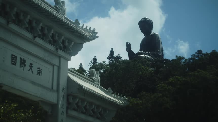 A chinese statue inside a temple at midday partially visible behind an entrance wall inscribed with chinese characters the scene is surrounded by lush trees with sunlight filtering through the foliage and casting soft shadows across the building