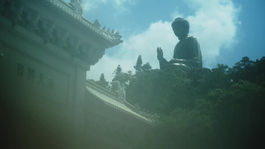 A buddha statue next to a building temple surrounded by plants under a cloudy sky in a sunny day