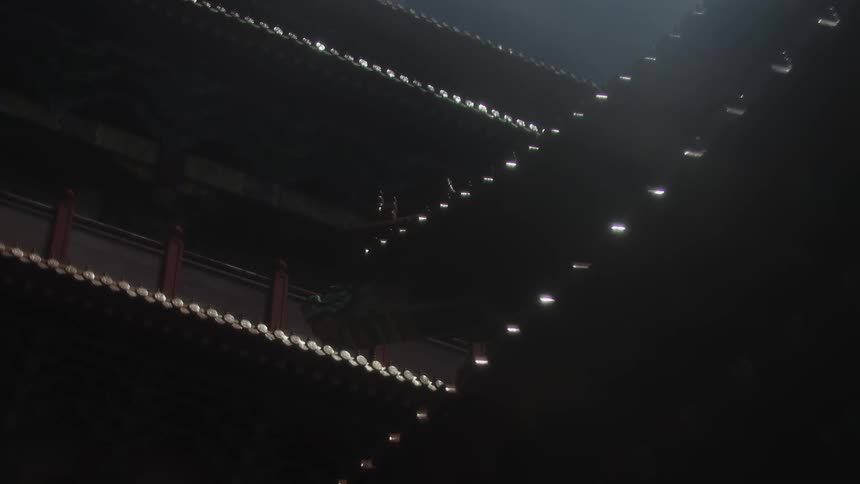 Rain drips down in shimmering vertical streaks from the tiled traditional chinese roof of a dark building with small lights visible on the eaves