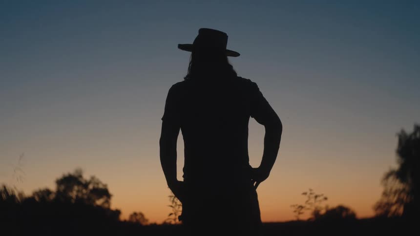 Silhouette of a young man wearing a hat standing outdoors at sunset