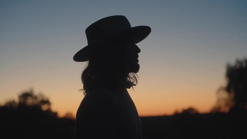 Female silhouette with widebrimmed hat stands before warmtoned sky at sunset
