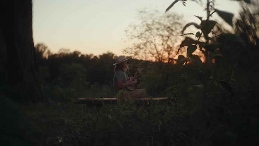 A young woman wearing a hat sitting on a bench in the park at sunset playing guitar as warm light filters through the trees