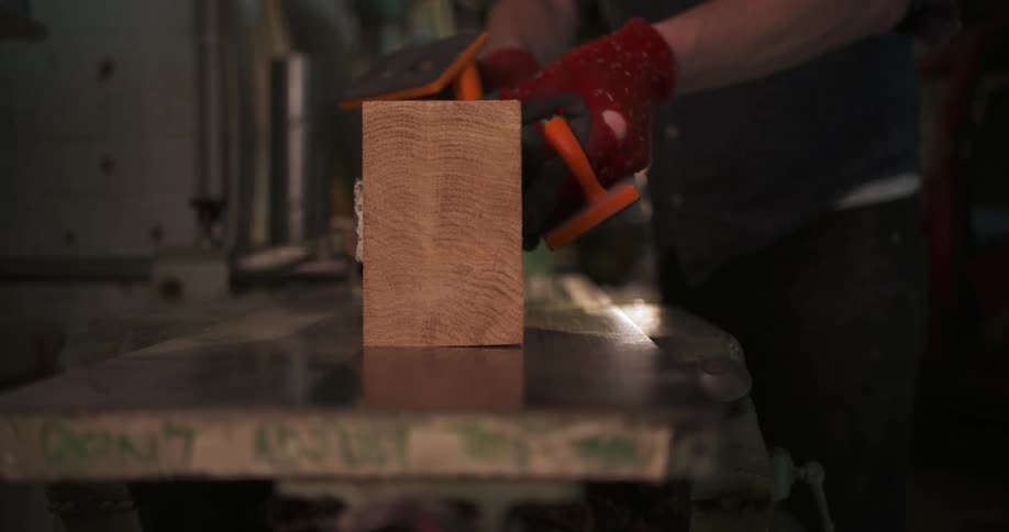 A worker in a woodshop is carefully pushing a large wooden block through a saw they are wearing red gloves and a darkcolored apron